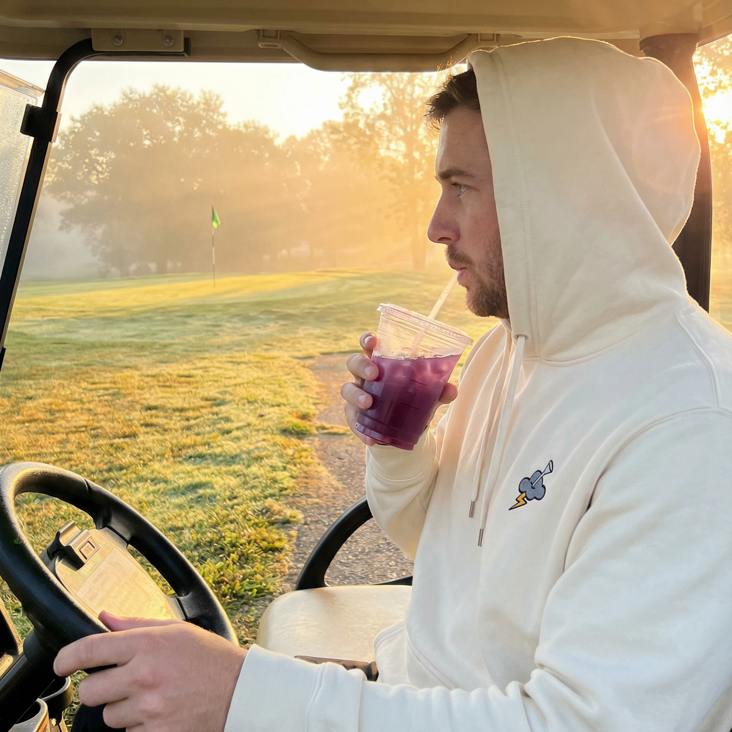 Man in a white hoodie sitting in a golf cart holding a drink on a golf course.