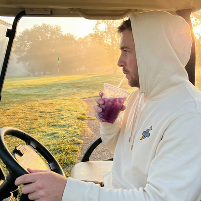 Man in a white hoodie sitting in a golf cart holding a drink on a golf course.